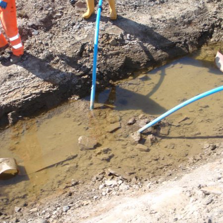 Construction worker standing at the edge of a muddy, partially-filled trench with a blue hose.